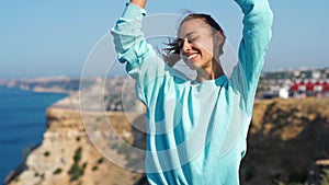 Portrait of happy young woman standing on cliff edge against amazing seascape and sky, hair blowing in the wind