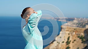 Portrait of happy young woman standing on cliff edge against amazing seascape and sky, hair blowing in the wind