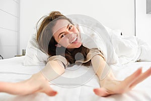 Portrait of happy young woman waking up energetic in morning, stretching arms towards camera and laughing in her bed