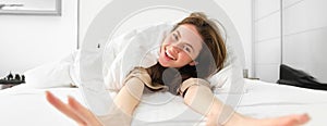 Portrait of happy young woman waking up energetic in morning, stretching arms towards camera and laughing in her bed