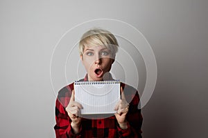 Portrait of hipster blonde woman posing in a red big cage shirt holding white empty notepad and poiting hang.Mock-up.