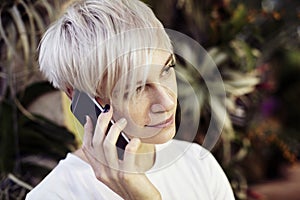 Portrait of hipster caucasian woman with blonde short hair talking by mobile phone. Smiling half-face face, Indoor botanical garde