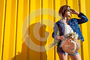 Portrait of hipster girl wearing glasses and hat with flowers against yellow background. Summer outfit. Fashion. Space.