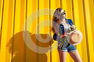 Portrait of hipster girl wearing glasses and hat with flowers against yellow background. Summer outfit. Fashion. Space