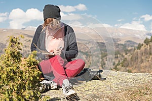 Portrait of a hipster girl wearing sunglasses and a hat sitting on a rock outdoor in the mountains against a blue sky