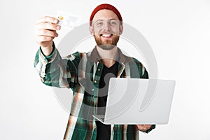 Portrait of hipster man holding silver laptop and credit card, while standing isolated over white background