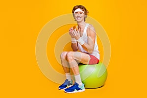 Portrait of his he nice attractive funky cheerful cheery sportive guy sitting on fitball eating forbidden tasty yummy