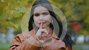 Portrait hispanic woman stands outdoors serious girl looking at camera holds index finger near mouth forbids speaking