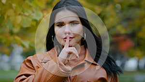 Portrait hispanic woman stands outdoors serious girl looking at camera holds index finger near mouth forbids speaking