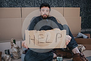 Portrait of homeless guy sitting on cardboard and holding a help cardboard in hands. He is looking straight at camera