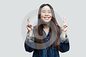 Portrait of hopeful beautiful brunette asian young woman in casual blue denim jacket with makeup standing with crossed finger and