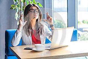 Portrait of hopeful beautiful stylish brunette young woman in glasses sitting, worried, closed eyes, crossed finger and hope, wish