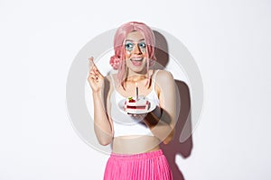 Portrait of hopeful birthday girl in pink wig, making wish with fingers crossed, holding b-day cake, standing over white