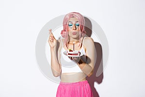 Portrait of hopeful birthday girl in pink wig, making wish with fingers crossed, holding b-day cake, standing over white