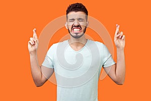 Portrait of hopeful brunette man crossing his fingers and eagerly wishing for good luck. indoor, isolated on orange background