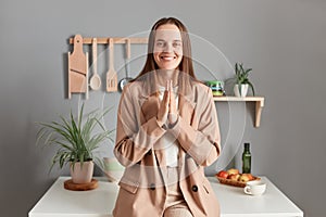 Portrait of hopeful grateful brown haired woman wearing beige suit standing near table on kitchen at home, looking at camera with