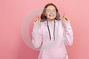 Portrait of hopeful joyous young woman with curly hair in hoodie raising fingers crossed while making wish, confident to win