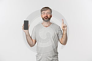 Portrait of hopeful man in gray t-shirt, showing mobile phone screen and making wish, crossing fingers for good luck