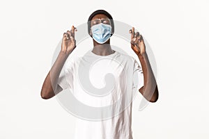 Portrait of hopeful nervous african-american man in beanie and medical mask, cross fingers good luck and making wish