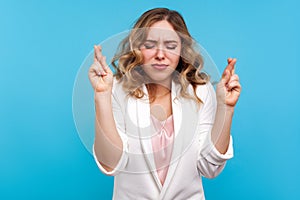 Portrait of hopeful woman standing with eyes closed and fingers crossed for good luck. blue background