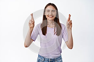 Portrait of hopeful young woman praying, cross fingers good luck and smiling with closed eyes, making wish, anticipating