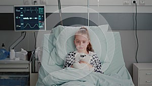 Portrait of hospitalized sick girl child patient holding teddy bear resting in bed during medical consultation