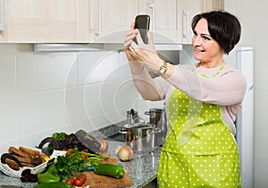 Portrait of housewife in apron making selfie in domestic kitchen