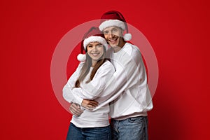 Portrait Of Hugging Young Couple In Santa Hats Posing Over Red Background