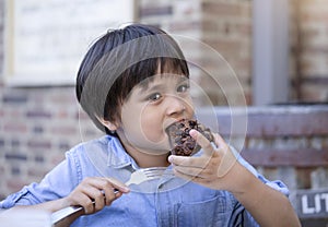 Portrait hugry little boy enjoy eating chocolate cake in outdoors cafe with blurry background of people, KId eating snack after pl
