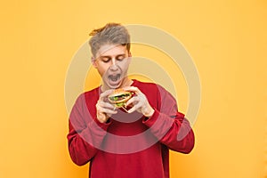 Portrait of a hungry boy with a burger in his hands on a yellow background. Young man with an open mouth looks at a burger holding