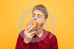 Portrait of a hungry guy in casual clothing bites an appetizing burger on a yellow background. Young man eats a burger, isolated.