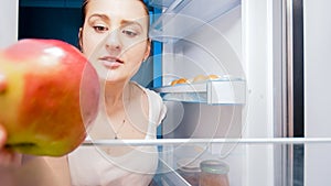 Portrait of hungry woman taking red apple out of fridge on kitchen at night. Concept of dieting and healthy nutrition