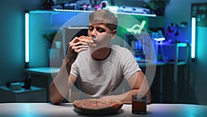 Portrait of hungry young man eating slice of tasty pizza with appetite sitting at table with drink on background of