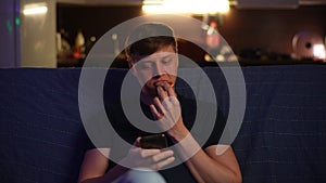 Portrait of hungry young man relaxing sitting on sofa in dark living room typing with friends on smartphone while eating