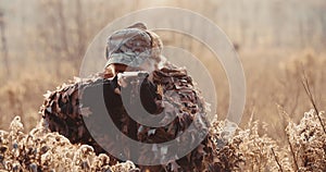 Portrait of hunter in hunting equipment aims with shot gun, lies in wait in the field in sunset light