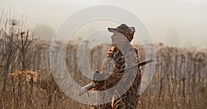 Portrait of hunter with shot gun standing in the field and looking around, foggy autumn morning, soft sun light