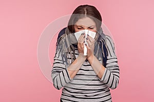 Portrait of ill woman wrapped in warm scarf sneezing in napkin, cleaning runny nose, suffering allergy