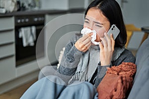 Portrait of ill young korean woman feeling sick, sneezing and holding napkin, staying at home ill, caught cold. Talking