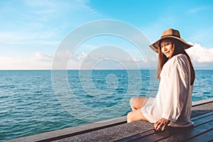 Portrait image of a happy beautiful asian woman on white dress sitting on wooden balcony by the sea with clear blue sky