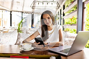 Portrait image of a young woman holding and using mobile phone and laptop in cafe