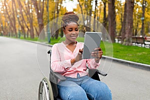Portrait of impaired young black woman in wheelchair using tablet pc for remote communication at autumn park