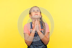 Portrait of imploring little girl in denim overalls looking up praying to god with pleading eyes, asking heartily
