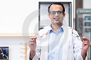 Portrait of Indian middle aged ophthalmologist or optometrist man work at optometry clinic, holding two different material and