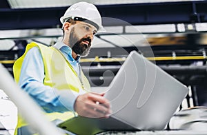 A portrait of an industrial man engineer with laptop in a factory, working.