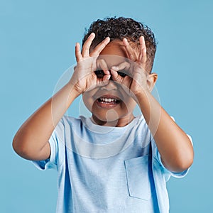 Portrait of inquisitive nosy little boy looking through fingers shaped like binoculars against a blue studio background