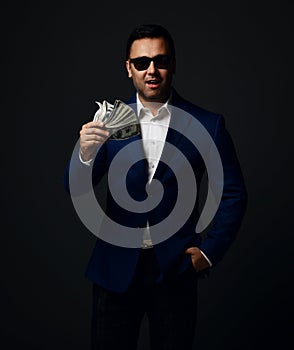 Portrait of insolent brunet businessman in blue jacket and sunglasses standing with fan of dollars cash in hand photo