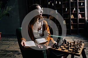 Portrait of intelligent young woman wearing elegant eyeglasses reading book sitting in armchair in dark library room, on