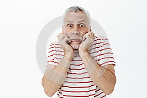 Portrait of intense anxious and scared charismatic senior man with grey beard and hair holding hands near mouth pursing