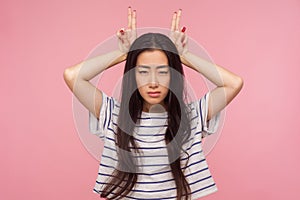 Portrait of irritated girl in striped t-shirt threatening with bull horn gesture, looking at camera with hostile expression