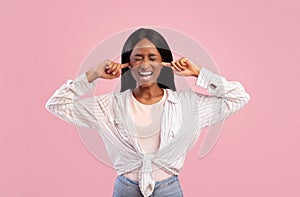 Portrait of irritated young black woman closing ears with fingers and shutting her eyes on pink studio background